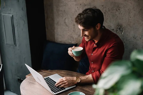 Sonriente joven freelancer trabajando en el ordenador portátil y beber café en la cafetería - foto de stock