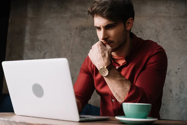 Pensativo joven freelancer trabajando en el ordenador portátil y beber café en la cafetería - foto de stock