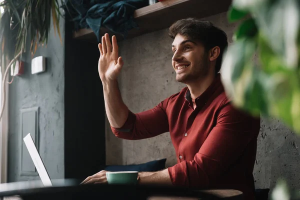 Sonriente joven freelancer trabajando en el ordenador portátil y agitando la mano en la cafetería - foto de stock