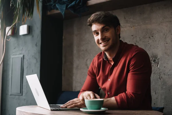 Souriant jeune pigiste travaillant sur ordinateur portable et boire du café dans un café — Photo de stock