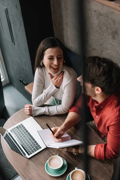 Enfoque selectivo de la joven pareja feliz trabajando en el ordenador portátil y riendo en la cafetería - foto de stock