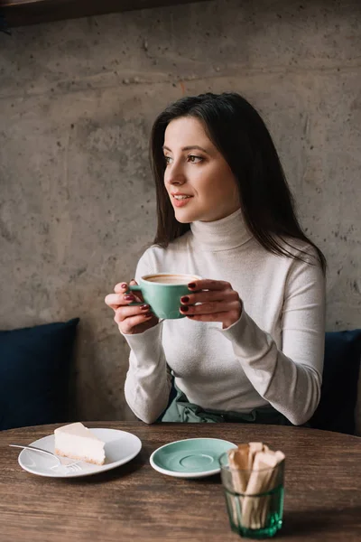 Улыбающаяся женщина пьет кофе и смотрит вдаль возле чизкейка в кафе — стоковое фото