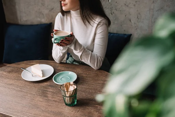 Messa a fuoco selettiva di pianta verde e donna che beve caffè vicino cheesecake in caffè — Foto stock