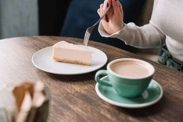 Vista parcial de la mujer comiendo pastel de queso con tenedor y beber café en la cafetería - foto de stock
