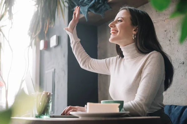 Selective focus of green plants and happy freelancer using laptop and waving hand in coffee shop — Stock Photo