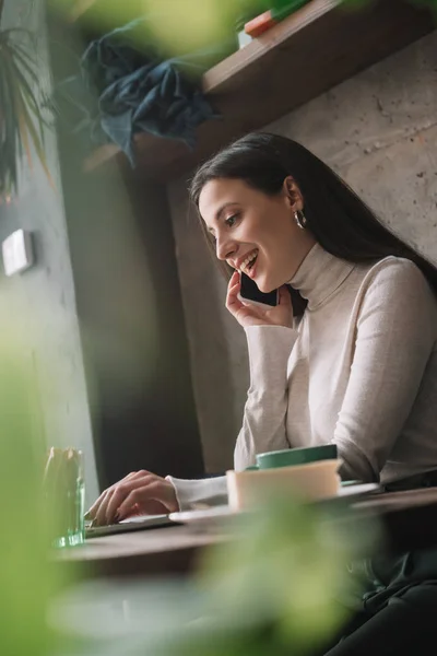 Selective focus of green plants and happy freelancer using laptop and talking on smartphone in coffee shop — Stock Photo