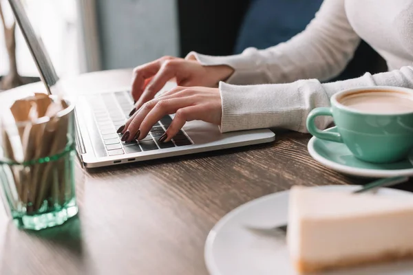 Vista recortada de freelancer utilizando el ordenador portátil cerca de café y pastel de queso - foto de stock