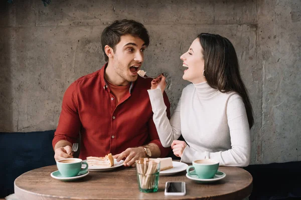 Happy girlfriend feeding boyfriend with cheesecake in coffee shop — Stock Photo