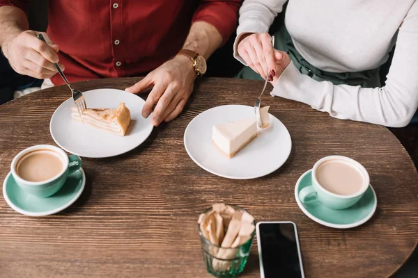 Cropped view of couple eating cheesecake and drinking cappuccino at wooden table with smartphone — Stock Photo