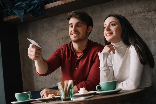 Pareja joven comiendo tarta de queso y tomando café mientras el hombre sostiene la tarjeta de crédito en la cafetería - foto de stock