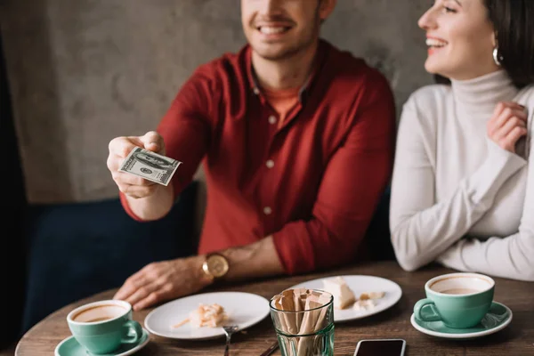 Cropped view of young couple eating cheesecake and drinking coffee while man holding money in coffee shop — Stock Photo