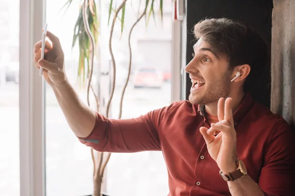 Людина в бездротових навушниках, використовуючи відео чат на смартфоні і показуючи знак миру біля вікна — стокове фото