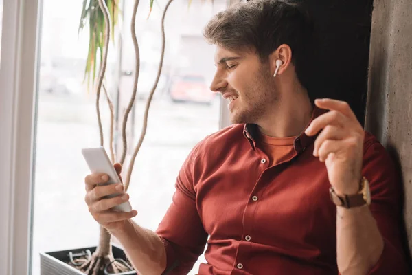 Sonriente hombre escuchando música en auriculares inalámbricos y utilizando smartphone teléfono inteligente cerca de la ventana - foto de stock