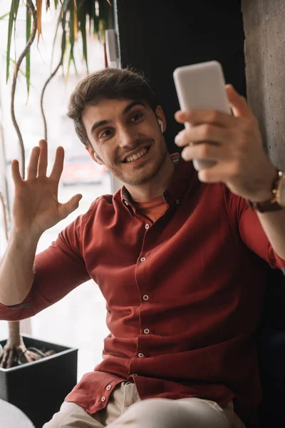 Hombre sonriente en auriculares inalámbricos utilizando tener chat de vídeo en el teléfono inteligente y agitar la mano cerca de la ventana - foto de stock