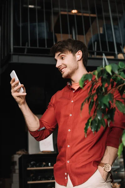 Uomo sorridente in auricolari senza fili utilizzando smartphone vicino pianta verde — Foto stock