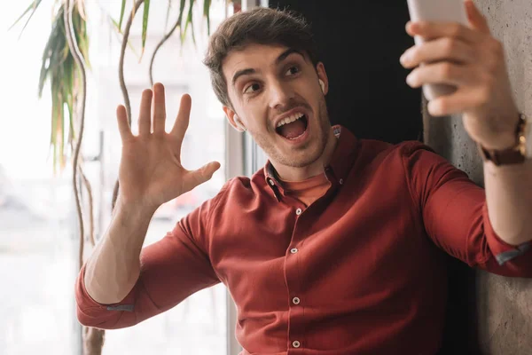 Mann mit drahtlosen Kopfhörern nutzt Videochat auf Smartphone und winkt mit der Hand am Fenster — Stockfoto