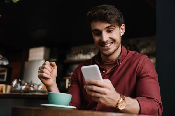 Happy man in wireless earphones using smartphone at table with cup of coffee — Stock Photo