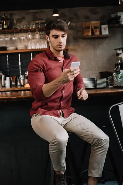 Hombre guapo sentado cerca del mostrador de bar en auriculares inalámbricos con teléfono inteligente - foto de stock