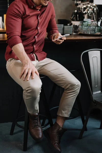 Обрезанный вид улыбающегося красивого мужчины, сидящего возле барной стойки со смартфоном — стоковое фото