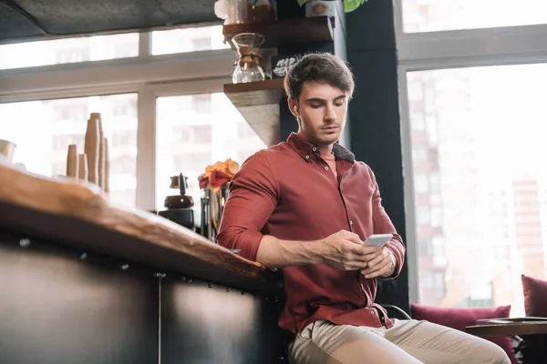 Hombre guapo sentado cerca del mostrador de bar en auriculares inalámbricos con teléfono inteligente - foto de stock