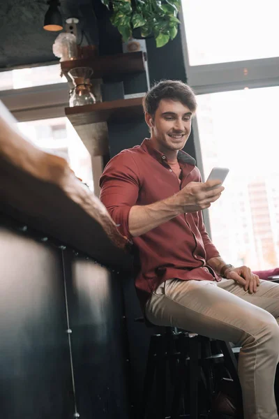 Hombre guapo sonriente sentado cerca del mostrador de bar en auriculares inalámbricos con teléfono inteligente - foto de stock