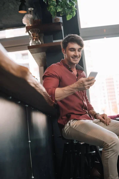 Smiling handsome man sitting near bar counter in wireless earphones using smartphone — Stock Photo