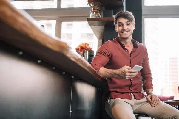 Sorrindo homem bonito sentado perto de bar contador em fones de ouvido sem fio usando smartphone — Fotografia de Stock
