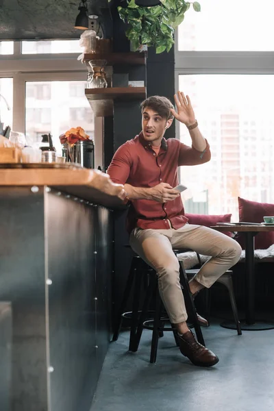 Schöner Mann sitzt in drahtlosen Kopfhörern mit Smartphone neben der Theke und winkt mit der Hand — Stockfoto