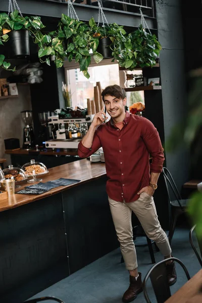 Smiling handsome man walking near bar counter and talking on smartphone — Stock Photo