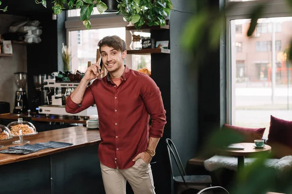 Sonriente hombre guapo caminando cerca del mostrador de bar y hablando en el teléfono inteligente - foto de stock
