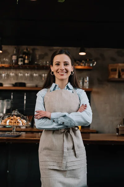 Smiling barista in apron standing with crossed arms near bar counter — Stock Photo