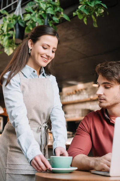 Cameriera sorridente che serve caffè a freelance con computer portatile — Foto stock