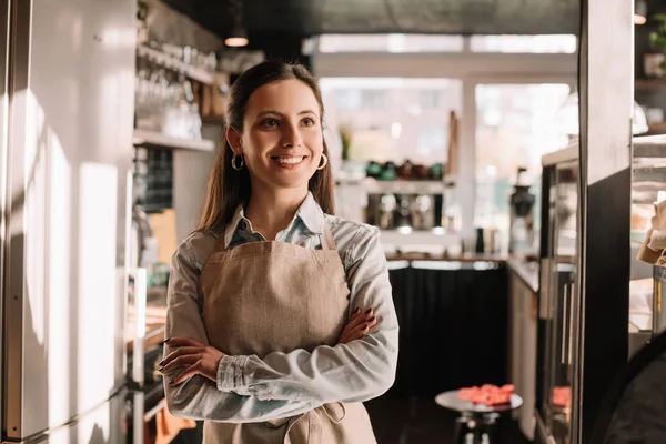 Sorridente barista in grembiule in piedi in soleggiato caffè con braccia incrociate — Foto stock