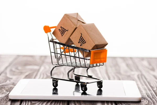 Carrito de compras de juguete con cajas de cartón pequeñas en tableta digital aislada en blanco, concepto de comercio electrónico - foto de stock