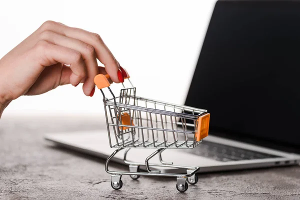 Vista cortada da mulher segurando carrinho de compras de brinquedo perto de laptop isolado no conceito branco, e-commerce — Fotografia de Stock