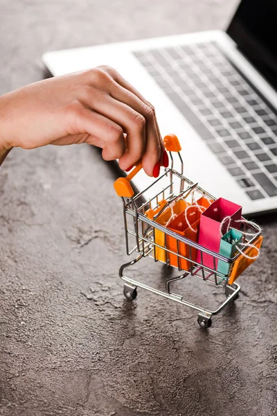 Cropped view of woman holding toy shopping cart with small shopping bags near laptop, e-commerce concept — Stock Photo