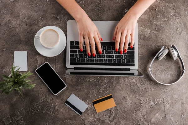 Vista superior de la mujer usando el ordenador portátil cerca de teléfono inteligente, taza de café, planta, auriculares y tarjetas de crédito, concepto de comercio electrónico - foto de stock