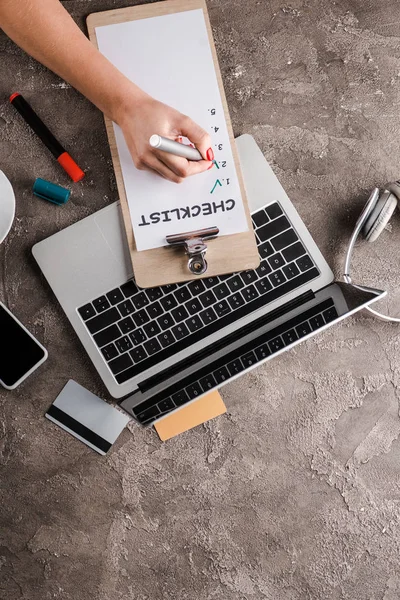 Vista superior de la mujer que sostiene la pluma marcador cerca de la lista de verificación, gadgets, tarjetas de crédito y auriculares, concepto de comercio electrónico - foto de stock