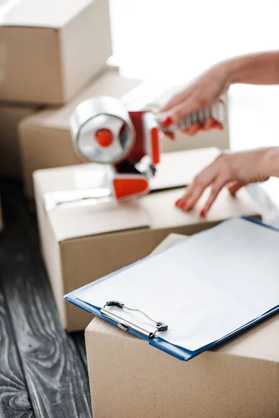 Cropped view of woman holding adhesive tape near carton box and clipboard in office — Stock Photo