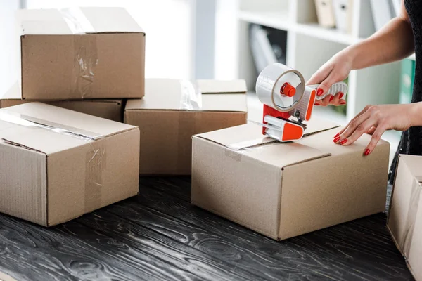 Cropped view of woman holding adhesive tape near carton box in office — Stock Photo