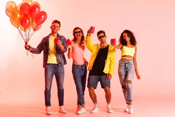 Happy multicultural friends holding plastic cups and balloons on pink — Stock Photo