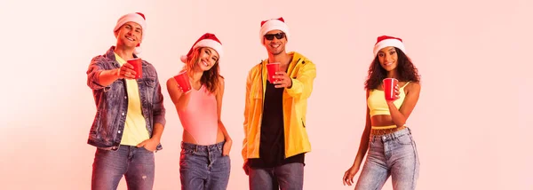 Panoramic shot of happy multicultural friends in santa hats holding plastic cups on pink — Stock Photo