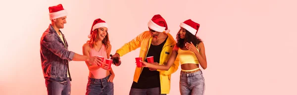 Panoramic shot of man in sunglasses pouring champagne in plastic cup near multicultural friends on pink — Stock Photo