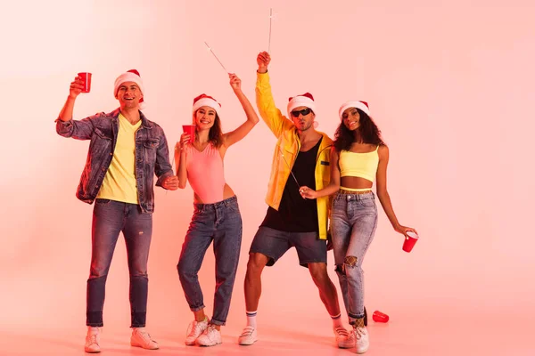 Happy multicultural fiends holding sparklers and plastic cups on pink — Stock Photo