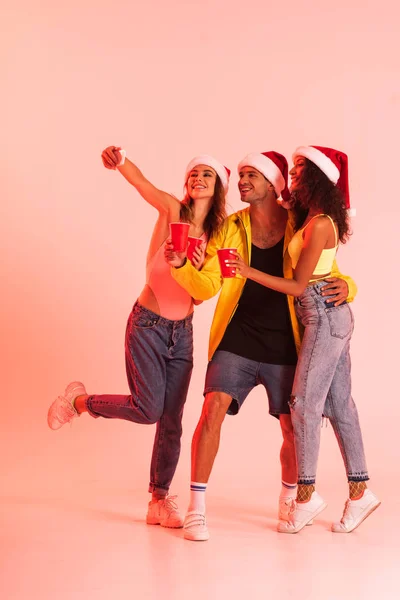 Felice ragazza prendendo selfie con amici multiculturali in cappelli di Babbo Natale su rosa — Foto stock
