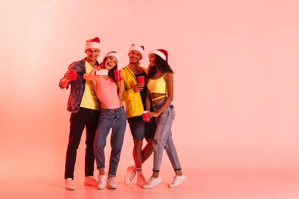 Femme heureuse prenant selfie avec des amis multiculturels dans chapeaux Santa sur rose — Photo de stock