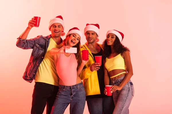 Alegre mujer tomando selfie con multicultural amigos en santa sombreros aislado en rosa - foto de stock