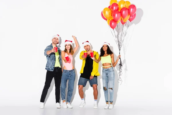 Felici amici multiculturali in cappelli di Babbo Natale con coppe di plastica vicino palloncini su bianco — Foto stock