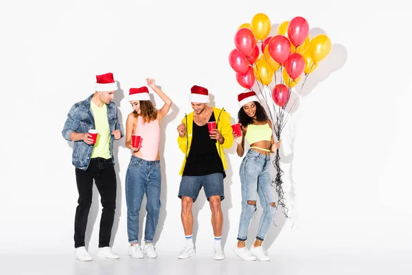 Cheerful multicultural friends in santa hats with plastic cups near balloons on white — Stock Photo