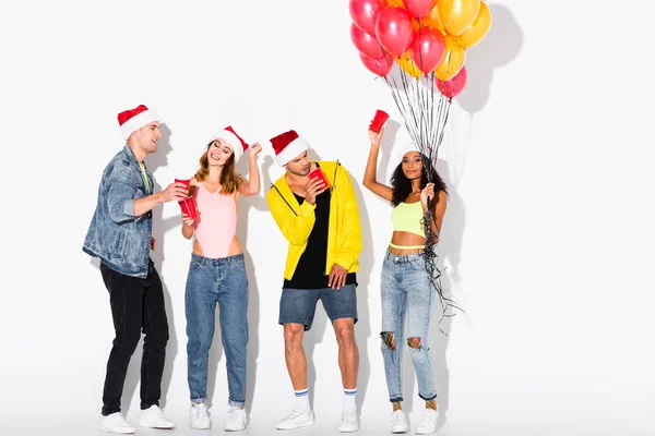 Uomini allegri e ragazze multiculturali in cappelli di Babbo Natale con coppe di plastica vicino palloncini su bianco — Foto stock